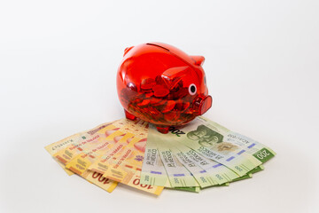 Red piggy bank full of Mexican coins perched on 100 and 200 peso bills.