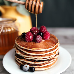 Juicy pancakes with berries and honey on a white plate, spoon, jar, wooden table