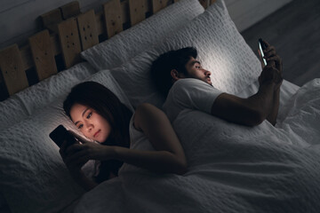 Young couple conflict in bed. happy smiling woman turned her back to man, reading message on mobile phone, trying to peek at screen. Cheating and infidelity concept.