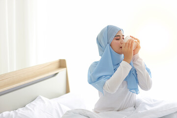 Beautiful and healthy asian muslim woman sitting on bed and drinking coffee or tea after getting up in the morning at sunrise in bedroom with happy and smiling face.