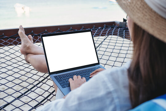Mockup image of a woman using and typing on laptop computer with blank desktop screen while lying down on hammock on the beach