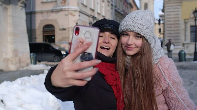 Two young smiling women tourists bloggers taking selfie photos portrait, video conferencing call on mobile phone on city street. Happy lesbian LGBT couple embracing. Winter holidays traveling vacation