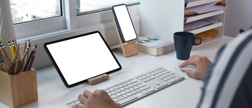 Horizontal image of young man working online with multiple devices at home office.