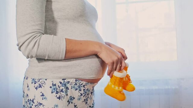 A young pregnant woman stands against a light window, holding knitted baby booties and walking them on her tummy.Belly and hands close-up. Concept of pregnancy and motherhood