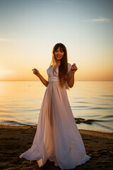 Fototapeta na wymiar Beautiful happy young woman in a white long dress on the seashore at sunset stands