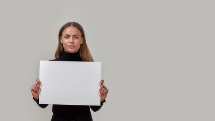 Portrait of young caucasian woman looking at camera, holding white blank banner in front of her while standing isolated over gray background