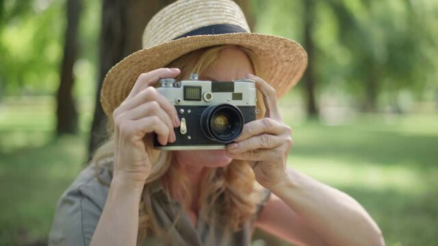 Close-up of camera with blurred middle aged woman taking photos of nature in summer park. Unrecognizable creative Caucasian lady enjoying hobby outdoors on sunny day.