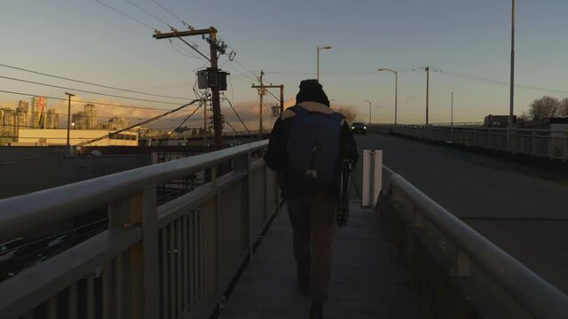 Following a travelling man walking along the sidewalk with guardrails on a bridge in the city while cars pass by in the evening with mountains in the background vancouver bc