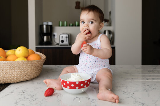 Cute baby sitting on kitchen counter eating strawberries with cream