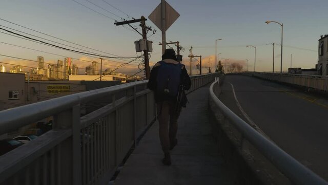Following a travelling man walking along the sidewalk towards a bridge in the city while cars pass by in the evening
