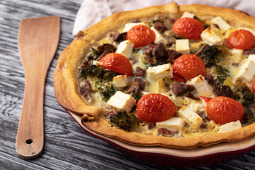 Quiche with meat, broccoli, tomato and brined cheese. On a black wooden table.
