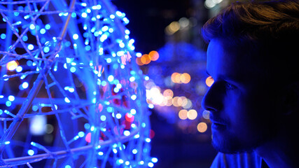 Side view of young millennial handsome man standing outside at night with lights and street out of focus in background