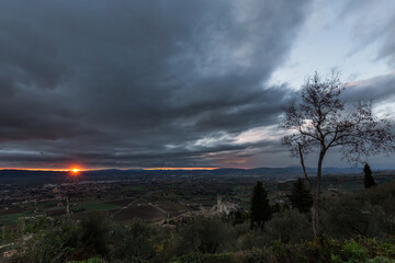 Distant sunset at Assisi town and St.Francis church with a moody sky and skeletal tree on the foreground