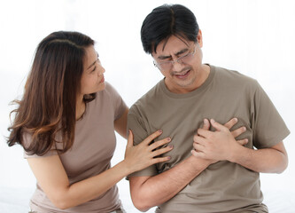 Portrait shot of middle-aged Asian lover couple wearing eyeglasses. Husband abruptly feels uncomfortable from the cardiac chest pain with concerned wife looking and supporting him with care.