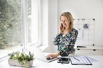 Businesswoman telephoning customer using cellphone while working at ergonomic standing desk in...