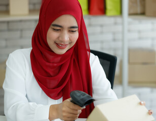 Portrait shot of cute smiling young teenage Muslim woman wearing red hijab sitting on a chair and scanning boxes with barcode scanner prepared for delivery. Concept of a modern online marketing
