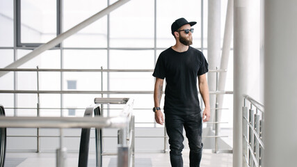 Hipster handsome male model with beard wearing black blank t-shirt and a baseball cap with space for your logo or design in casual urban style