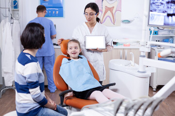 Obraz premium Dentist in clinic with chroma key on tablet pc display consulting kid with bad tooth. Stomatolog explaining teeth prevention to mother and child holding tablet pc with copy space available.