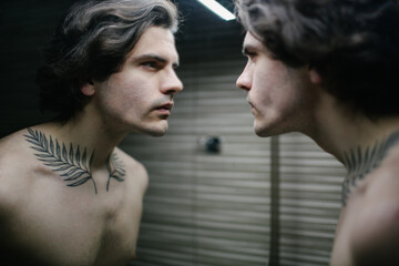 Young man in bathroom looking into a mirror at himself. He has a tattoo over his neck