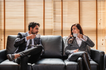 A businesswoman and a businessman in suits enjoying coffee together sitting on a black sofa by the window. Looking at each other.