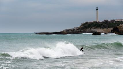 Surfeur isolé surfant une vague à Biarritz, France. Le phare de Biarritz est visible en arrière-plan. Ciel nuageux d'hiver
