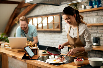 Young happy woman preparing fried eggs with bacon for breakfast in the kitchen.
