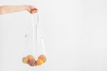 Hand holding white mesh bag with fruits on white background, copy space. Eco friendly, reusable shopping bag. Oranges, apples, garnet in cotton knitted string bag. Zero waste concept.
