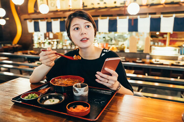 Asian chinese young woman enjoy japanese food rice with eel grilled and scallop in bowl in fish market tokyo japan. female tourist sitting in local traditional izakaya trying unadon using cellphone