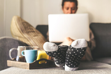 Woman sitting on a couch in the living room with warm socks in a winter morning. Girl using laptop...