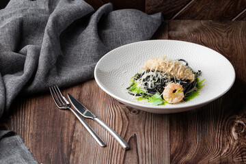 Black spaghetti. Black seafood pasta with shrimps and parmesan on white plate isolated on wooden background. Silver cutlery and grey linen napkin by side. Mediterranean gourmet food.