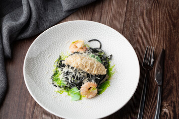 Black spaghetti. Black seafood pasta with shrimps and parmesan on white plate isolated on wooden background. Silver cutlery and grey linen napkin by side. Mediterranean gourmet food.