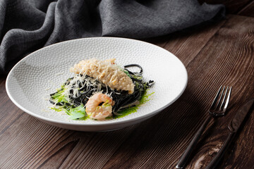 Black spaghetti. Black seafood pasta with shrimps and parmesan on white plate isolated on wooden background. Silver cutlery and grey linen napkin by side. Mediterranean gourmet food.