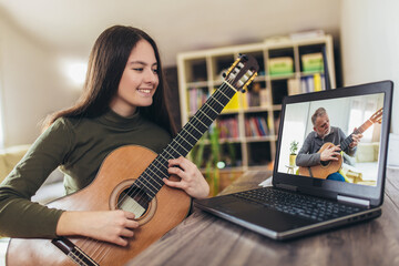 Focused girl playing acoustic guitar and watching online course on laptop while practicing at home....