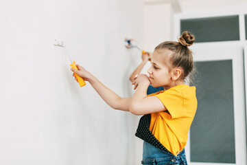 A girl in a denim overalls and a yellow T-shirt helps to paint the walls in an apartment white