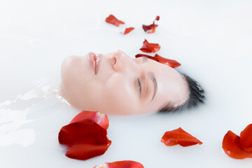 Close up female face in the milk bath with soft white glowing and rose petals. Copyspace for advertising. Beauty, fashion, style, bodycare concept. Attractive caucasian model in milky colored foam.