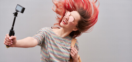 Young blogger woman with pink dyed flying hairs dancing near the grey wall and making vlog by...
