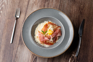 Pasta Carbonara. Spaghetti with bacon, parmesan, cream sauce and egg yolk. Isolated dish on a grey round plate with silver cutlery on wooden background. Mediterranean gourmet food. Top view.