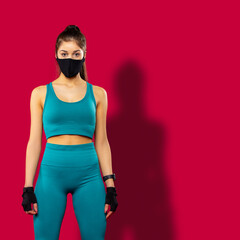 Fototapeta na wymiar Brunette sportwoman wearing blue fitness clothing and black medical protective face mask training indoors. Fitness fit form, health and body care during covid-19 on red background
