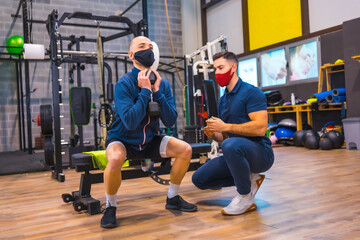 Fototapeta na wymiar Instructor in the gym correcting the squats of the young athlete in the coronavirus pandemic, a new normal. With protective face mask