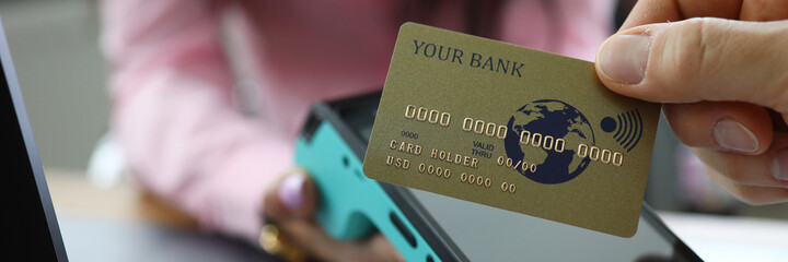 Man make cashless payment. Gold card is attached to terminal. Woman hold terminal in her hand and give it to man for payment. Banking operation through payment instrument. There is gray laptop on