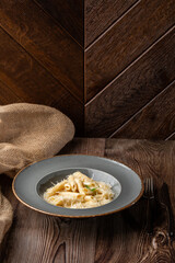 Pasta Penne with parmesan cheese and creamy sauce. Isolated dish in a grey round plate on wooden background. Sacking and silver cutlery by side. Mediterranean gourmet food. Copy space.