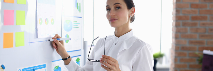 Beautiful woman in white blouse hold glasses in one hand and draw chart on board with other hand. The most modern presentation tools: interactive whiteboard, system, complexe. Employee show and draw