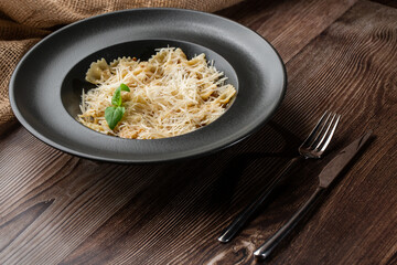 Pasta Farfalle with parmesan cheese and creamy sauce. Isolated dish in a black round plate on wooden background. Sacking and silver cutlery by side. Mediterranean gourmet food.