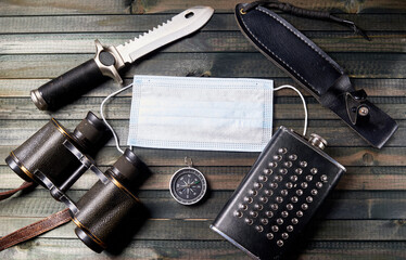 Up-to-date travel kit: medical mask, compass, binoculars, flask, tactical knife on dark wooden background