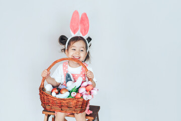 A little girl dressed as a rabbit holding a large basket filled with eggs and tulips against a white background