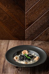 Black spaghetti. Close-up of black pasta with shrimps and parmesan in a black round plate isolated on wooden background. Mediterranean gourmet food. Vertical orientation, copy space.