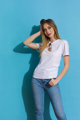 Young Blond Woman In White Shirt And Jeans Is Posing In The Sunlight Against Blue Wall.