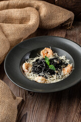Black spaghetti. Black pasta with shrimps and parmesan in a black round plate isolated on wooden background with sacking by side. Mediterranean gourmet food.
