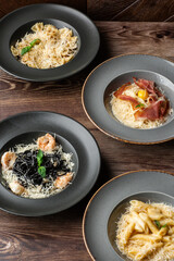 Four kinds of different cooked pastas: Penne, Farfalle, Carbonara, Black Spaghetti. Close-up isolated on wooden background in black and grey plates. Mediterranean gourmet food. Top view.