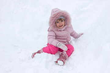 Fototapeta na wymiar Little girl in pink winter clothes stands on a snow-covered city street under a snowfall with joyful emotions on her face, happy childhood, active winter lifestyle
